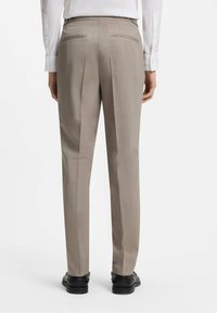 Homme portant un pantalon beige sur mesure, une chemise blanche et des chaussures noires, vu de dos sur fond blanc uni.