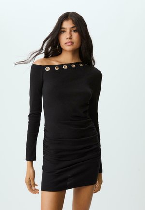 Eine Frau trägt ein schwarzes, einseitig geschnittenes Minikleid mit langen Ärmeln und metallischen Ösen am Ausschnitt, während sie vor einem schlichten Hintergrund steht.