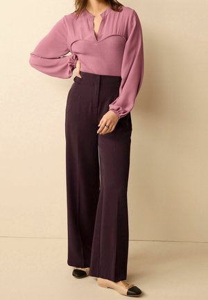 Femme debout avec la main sur la hanche, portant un chemisier mauve à manches longues, un pantalon large taille haute violet foncé, et des ballerines beiges à bouts noirs.
