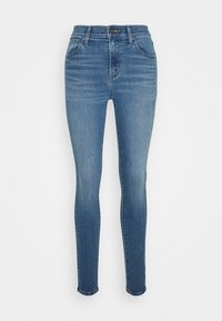 Blå tajta jeans i denim med framfickor, bältesöglor och knappstängning, uppvisade mot en vit bakgrund.
