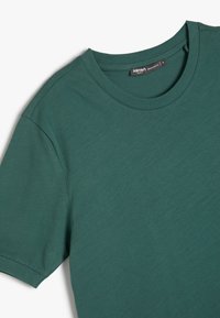 Baumwoll-T-Shirt in einem tiefen Grünton, mit Rundhalsausschnitt und kurzen Ärmeln. Weiche Textur mit einem einfachen, schlichten Design.