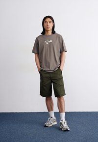 Ung mand med langt hår står selvsikkert i et minimalistisk rum, iført en grå t-shirt, olivenfarvede shorts og træningssko.