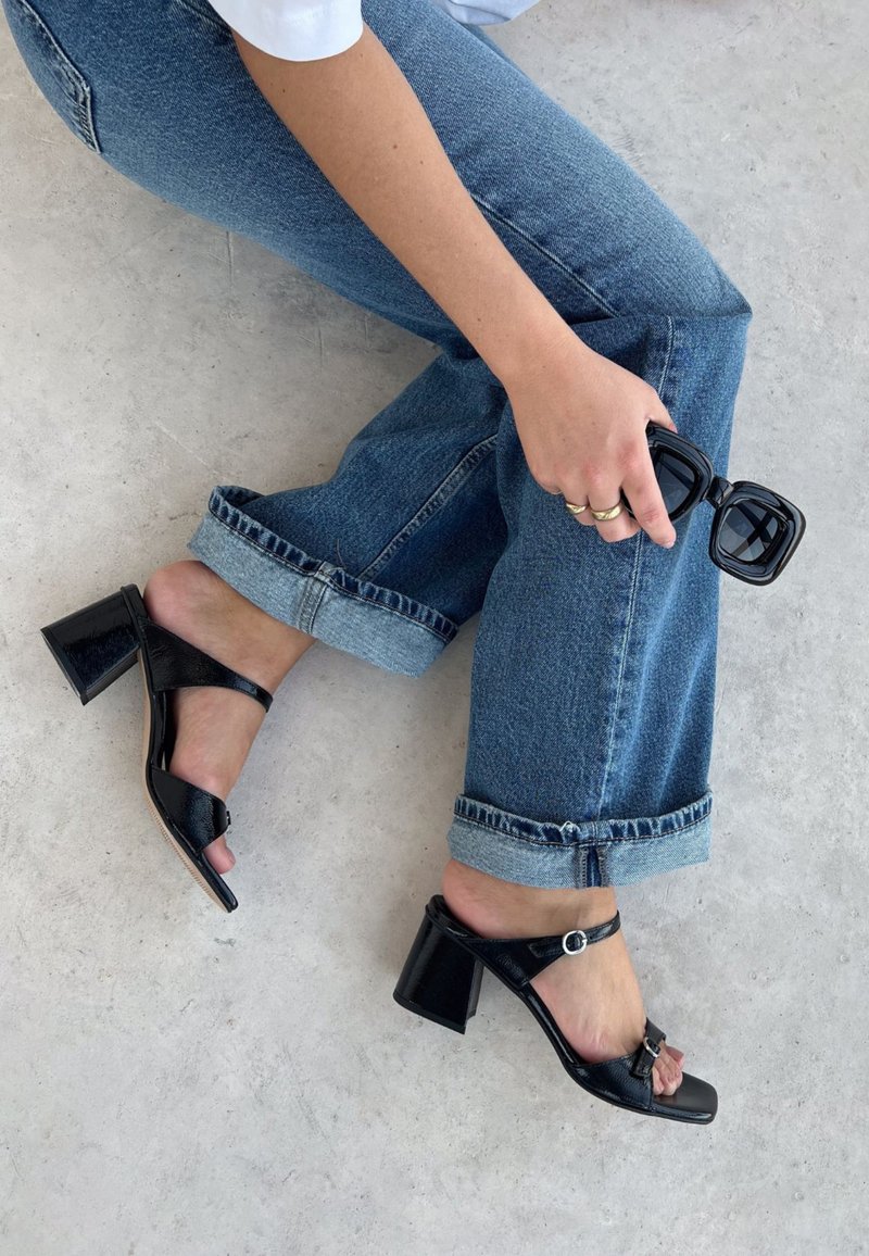 Sandales à talons bloc en cuir verni noir avec une large sangle, une boucle réglable à la cheville et un design à bout ouvert. Assorties avec un jean retroussé.