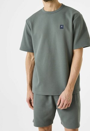Mand iført tekstureret grågrøn kortærmet skjorte og matchende shorts, stående mod en ensartet hvid baggrund.