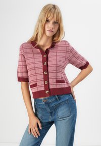 Femme aux cheveux blonds portant un cardigan en tricot à carreaux rose et bordeaux avec des boutons, et un jean bleu, sur fond blanc.