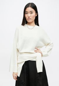 Sweater branco oversized com punhos e bainha canelados, apresentando um detalhe de cintura atada, combinado com uma saia preta adornada com bordados florais.