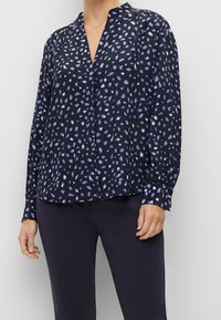 Navybluse mit V-Ausschnitt und langen Ärmeln, die ein verstreutes lila-weißes Muster aufweist. Hergestellt aus leichtem Stoff mit glatter Textur.