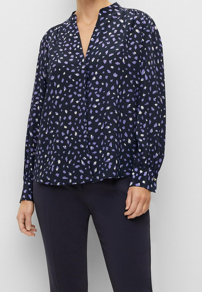 Navybluse mit V-Ausschnitt und langen Ärmeln, die ein verstreutes lila-weißes Muster aufweist. Hergestellt aus leichtem Stoff mit glatter Textur.