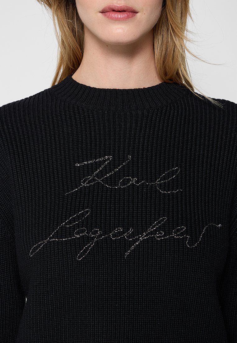Pull en maille noir avec tissu texturé, orné d'une broderie en cursive argentée indiquant "Karl Lagerfeld". Encolure ronde et détails côtelés.
