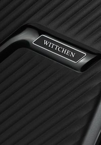 Black suitcase with vertical ribbed texture, featuring a glossy black handle and a metallic brand tag embossed with "Wittchen."