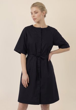 Frau mit blondem Haar in einem schwarzen knielangen Kleid mit halblangen Ärmeln und gebundenem Taillenband, stehend vor einem beigen Hintergrund.