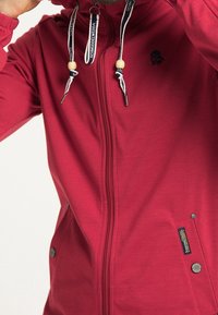 Veste à capuche zippée rouge avec une finition texturée, dotée d'une capuche avec cordons de serrage réglables, de poches latérales et de détails de logo brodé.