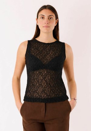 Donna con capelli lunghi e scuri che indossa una maglietta nera senza maniche in pizzo e pantaloni marroni, in piedi con le mani nelle tasche davanti a uno sfondo semplice.