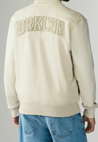 Felpa crema con lettering in rilievo "BRKLN" e "Williamsburg" sotto, con polsini e orlo a coste. Abbinata a jeans in denim blu.