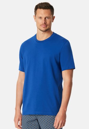 Mann trägt ein einfarbiges königsblaues T-Shirt mit kurzen Ärmeln und gemusterte blaue Shorts, stehend vor einem weißen Hintergrund.