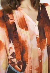 Blusa multicolore nelle tonalità arancione, marrone e rosa. Caratterizzata da un collo a V, chiusura frontale con bottoni e pieghe verticali sulle spalle. Tessuto morbido al tatto.