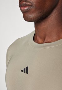 Beżowa koszulka sportowa z okrągłym dekoltem i czarnym logo na piersi, wykonana z gładkiego, elastycznego materiału.