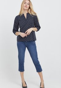 Blusa blu con motivo bianco, maniche a tre quarti e scollatura con bottoni, abbinata a jeans blu cropped e tacchi neri.