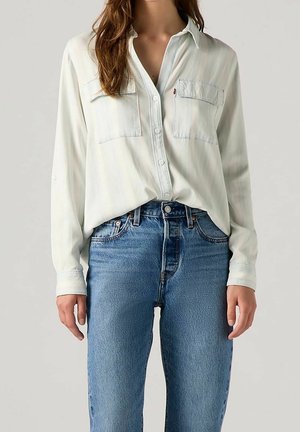 Mujer con una camisa blanca holgada abotonada con dos bolsillos en el pecho combinada con vaqueros de mezclilla azul de talle alto sobre un fondo liso.