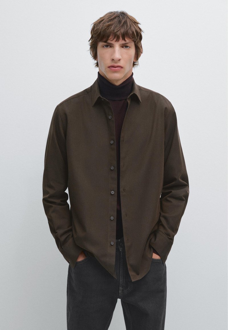 Camicia marrone con bottoni, dalla texture liscia e colletto standard, indossata sopra un collo alto scuro. Il modello sta in piedi con le mani in tasca contro uno sfondo chiaro.