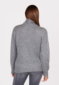 Vrouw met lang bruin haar, gekleed in een grijze ribgebreide coltrui en grijze spijkerbroek, van achteren bekeken tegen een witte achtergrond.