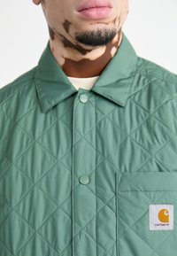 Chaqueta verde acolchada con costuras en forma de diamante, con un cuello abotonado, bolsillo frontal y una etiqueta con un logo.