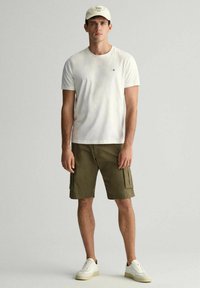 T-shirt bianco in cotone abbinato a pantaloncini cargo verdi oliva, dotati di tasche laterali. L'outfit è completato da sneaker bianche e un cappellino leggero.