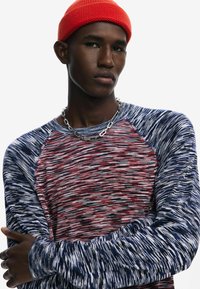 Punane ja tumesinine triibuline pikkade varrukatega särk karedate kudumismustritega, koos erkpunase beanie ja hõbedase kettkaelakeega.