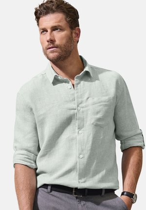 Man with short curly hair and beard wearing light green button-up shirt with sleeves rolled up and gray pants with black woven belt.