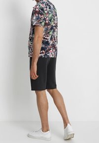 Muž oblečený v tričku s krátkým rukávem s tropickým potiskem, černých kraťasech po kolena a bílých teniskách, stojící u prosté zdi.