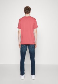 Camiseta de algodón roja con mangas cortas y escote redondo, combinada con jeans de mezclilla azul oscuro y zapatillas blancas, vista desde la parte trasera.