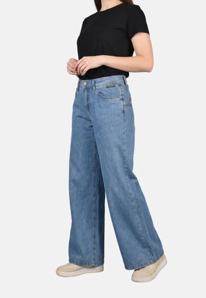 Mujer con jeans anchos de pierna en azul claro, camiseta negra de manga corta y zapatillas beige, de pie con las manos entrelazadas cerca de la cintura.