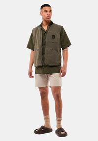 Gilet verde oliva con grandi tasche, indossato sopra una camicia scura a maniche corte e pantaloni corti crema. Sandali marroni e calzini beige visibili.