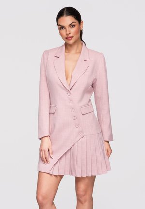 Femme aux cheveux foncés attachés en queue de cheval basse, portant une robe blazer asymétrique rose clair avec des boutons et une partie jupe plissée.