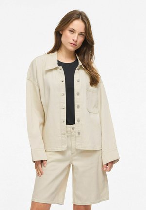 Pikkade juustega naine kannab musta topi peal beežikat nööbitavat jopet ja sobivaid beeže põlvikmuni pükse, seistes ühevärvilise tausta ees.