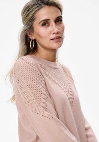 Kobieta z długimi blond włosami, nosząca różowy sweter o dzianinowym wzorze z teksturą oraz dużymi złotymi kolczykami w kształcie koła, na białym tle.