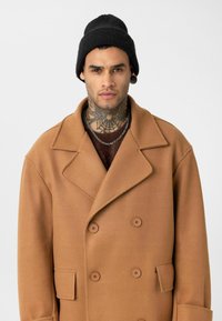 Jonge man met nektattoo en zwarte beanie draagt een camelkleurige, dubbelgeknoopte jas en gelaagde zilveren kettingen tegen een witte achtergrond.