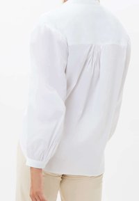 Blusa blanca con cuello alto, detalle de parte trasera fruncido y mangas largas voluminosas. El tejido parece suave y ligero.