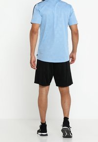 Man som står med ryggen mot oss, iförd en ljusblå mönstrad sportskjorta, svarta shorts och svarta träningsskor på en vit bakgrund.