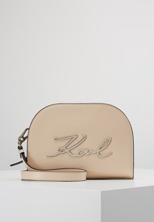 Borsa a tracolla in pelle beige con forma curva, caratterizzata da un logo in corsivo argentato "Karl". Include una tracolla regolabile e una superficie liscia.