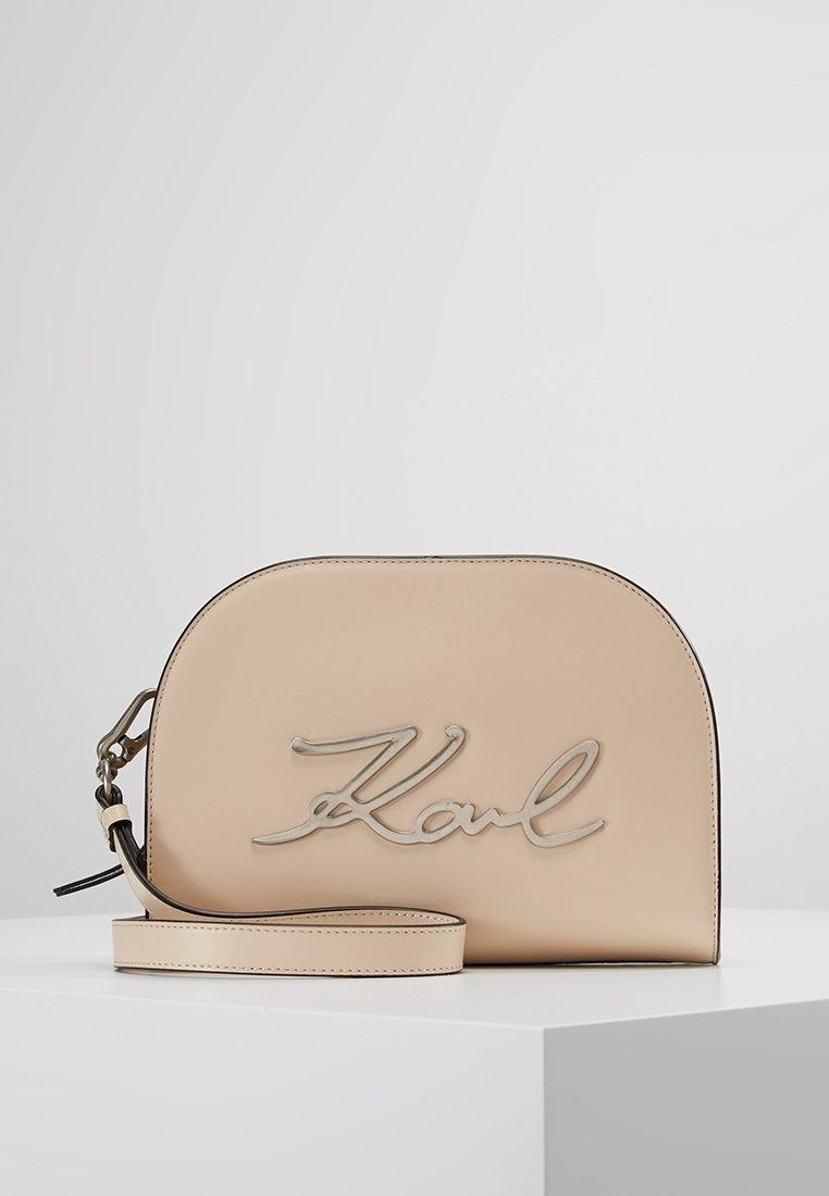 Beige läderhandväska med en böjd form, försedd med en silverfärgad kursiv "Karl"-logga. Inkluderar en justerbar rem och en slät yta.