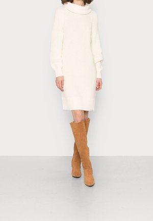 Person iført en knælængde hvid strikket kjole og brune knæhøje ruskindsstøvler, stående på en lys grå gulv mod en hvid væg.