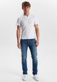 Hvid v-hals t-shirt, denim jeans med gradient fra lys til mørk blå og hvide sneakers. Modellen står oprejst foran en ensfarvet baggrund.