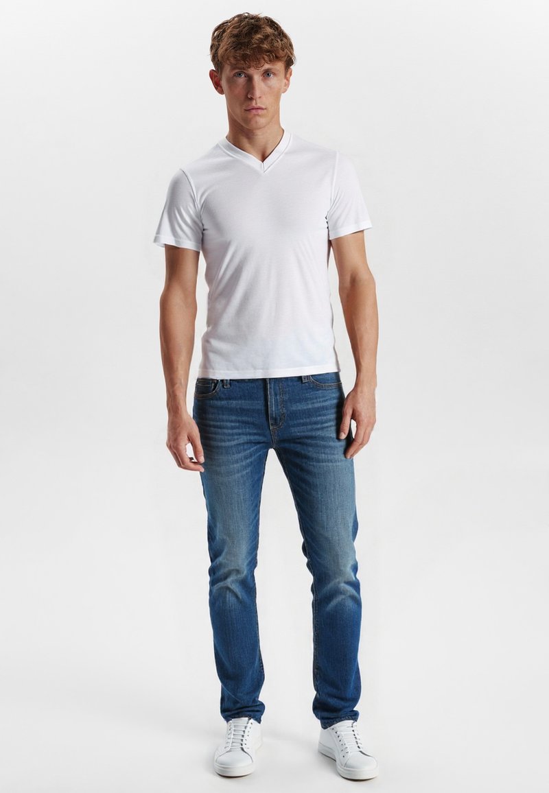 Hvid v-hals t-shirt, denim jeans med gradient fra lys til mørk blå og hvide sneakers. Modellen står oprejst foran en ensfarvet baggrund.