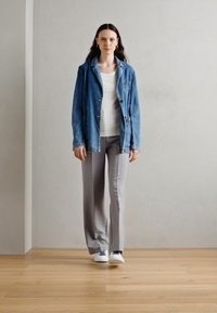 Jachetă din denim cu croială lejeră, cămașă albă pe dedesubt, pantaloni formali gri și adidași albi pe un podea din lemn, împotriva unui perete deschis la culoare.
