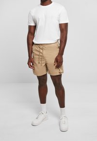 Shorts beiges avec une taille élastique et un cordon de serrage, dotés de poches cargo latérales, portés avec un t-shirt blanc et des baskets blanches.