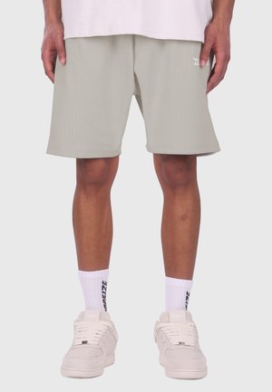 Personne portant un short côtelé gris clair, des chaussettes blanches avec un texte noir et des baskets blanc cassé, debout devant un fond uni.