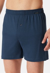 Marineblaue Boxershorts mit elastischem Bund, ausgestattet mit einem strukturierten geometrischen Muster und einem einzelnen Knopfverschluss an der Vorderseite.