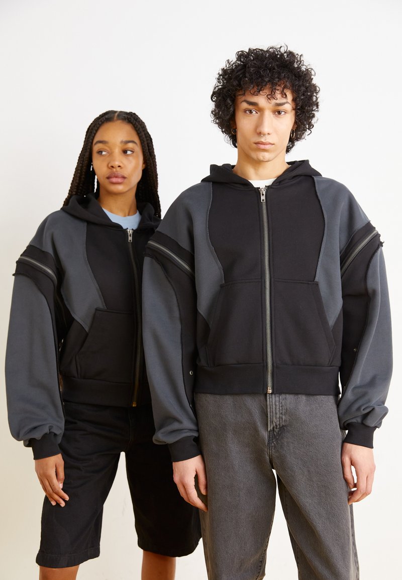 Black and gray zip-up hooded sweatshirts with contrasting panels, oversized fit, kangaroo pockets, and ribbed cuffs, worn over casual bottoms.