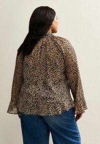 Kobieta stojąca plecami, ubrana w bluzkę w brązowo-czarne printy lamparta z szerokimi rękawami oraz niebieskie dżinsy, na jednolitym tle.